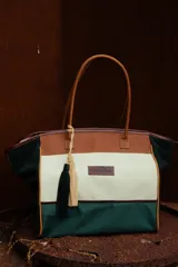 Bolso de fin de semana marca Sierra Mora, confeccionado en lona y gamuza a rayas horizontales en colores verde oscuro, blanco y marrón. Tiene doble asa de cuero marrón, cierre superior y bolsillo interno. Incluye dos borlas decorativas colgantes en verde y beige.