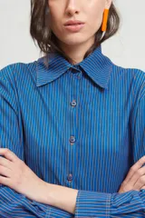 Camisa azul con rayas verticales color naranja, de corte holgado, mangas largas con pinzas laterales y puños anchos. Cuello clásico de gran tamaño.