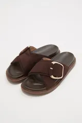 Sandalias tipo pantufla de color marrón oscuro, con tiras cruzadas de material tipo gamuza y hebilla grande de metal color oro rosa. Tienen una plataforma o suela gruesa de goma marrón.