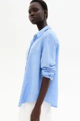 Camisa celeste de tejido liviano en mezcla de lino y viscosa, con cuello clásico, botones al frente, canesú de doble capa con pliegue de caja en la espalda, mangas largas con puños abotonados, bolsillo superior abierto y bajo redondeado.