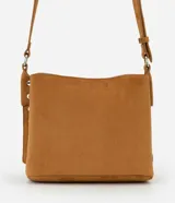 Cartera de gamuza color tostado, de tamaño mediano, con diseño transversal y asa larga ajustable. Cuenta con cierre superior de botón a presión, compartimento interno principal y detalles laterales con ojales metálicos.