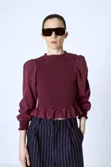 Blusa color borgoña con escote redondo con volantes, cuerpo fruncido, mangas largas abullonadas y puños elásticos con volantes.