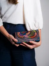 Billetera tipo sobre con cierre de cremallera, confeccionada artesanalmente en Tailandia. Presenta bordados multicolores con motivos de aves y flores. Incluye un tirador con borla y cuentas de madera.