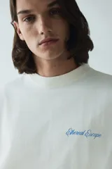 Remera color hueso de corte oversized, confeccionada en tejido de algodón pesado. Presenta cuello redondo acanalado, hombros caídos y estampado pequeño con la frase "Ethereal Escape" en el lado izquierdo del pecho.