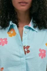 Camisa celeste con estampado de flores rosas, naranjas y marrones, de manga corta, cuello camisero y botones nacarados. Confeccionada en tejido seersucker.