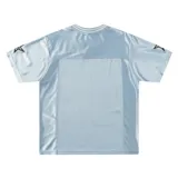 Remera estilo jersey de fútbol o americana, color celeste claro, con paneles de tela satinada y malla microperforada. Presenta cuello en V con ribete blanco, el número "90" grande en el frente con borde negro, y texto en la parte superior que dice "BAEN MADE" y "Madcapsclub".