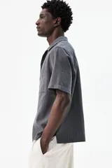 Camisa de manga corta color gris oscuro, confeccionada en mezcla de lino y algodón. Tiene cuello abierto, tapeta lisa, bolsillo de parche en el pecho y bajo recto con aberturas laterales.