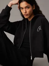 Buzo negro cropped con capucha y cierre frontal de cremallera metálica. Tiene bolsillos de parche en la cintura y logo de Calvin Klein bordado en el pecho.