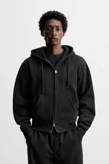 Sudadera negra relaxed fit de algodón compacto con interior en rizo, cuello con capucha ajustable, manga larga, bolsillos frontales tipo canguro, acabados en rib y cierre frontal con cremallera.
