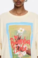 Camiseta color crema de algodón con estampado de un cuadro con borde celeste, con una mano sosteniendo una flor y el texto "Bimba y Lola".