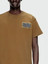 Remera de manga corta color marrón, con cuello redondo y un pequeño parche estampado con diseño abstracto en el pecho.