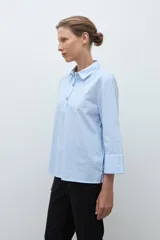 Camisa de popelina celeste con finas rayas blancas verticales, cuello camisero y abertura frontal con botones joya. Manga larga con puño ancho.