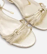 Sandalia plana marca Satinato, color dorado metalizado con textura tipo croco. Posee tiras finas con detalle de nudo en el empeine y cierre de lazo ajustable al tobillo. Diseño de puntera redonda y suela plana.