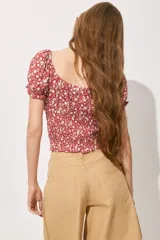 Blusa roja con estampado de flores blancas, escote cuadrado con cordón ajustable, mangas cortas abullonadas con elástico en hombros y puños, cintura fruncida y volados en mangas y bajo.
