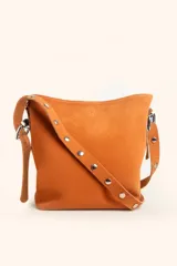 Cartera tipo balde de gamuza color suela, con correa ajustable decorada con tachas metálicas plateadas. Presenta un diseño minimalista con logo grabado en la parte superior frontal.