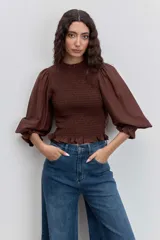Blusa color marrón con cuerpo fruncido tipo nido de abeja, cuello alto con volados y mangas largas abullonadas con puños elásticos terminados en volados.