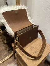 Bandolera de cuero color negro con forro interior y cierre. Presenta un cuerpo cubierto de lana de oveja color blanco y correa larga de cuero marrón con hebilla ajustable.