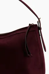 Bolso de hombro color borgoña, con textura que simula gamuza o ante, de forma curva y cierre superior con cremallera. Posee una correa de hombro delgada de cuero marrón oscuro y un tirador de cremallera con borla a juego.