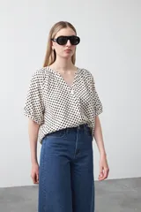 Camisa de viscosa con estampado de lunares pequeños en negro sobre fondo blanco o crema. Presenta escote en V con abertura y botones frontales, y mangas cortas abullonadas.