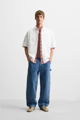 Camisa blanca de algodón con estructura canvas, corte boxy fit, cuello bowling y manga corta. Presenta bordado en azul claro con texto cursivo en el pecho y botones frontales.