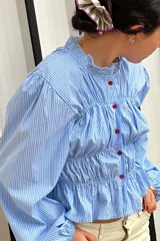 Camisa celeste a rayas verticales blancas, de corte entallado, con cuello clásico, cierre frontal con botones rojos, frunces en el delantero y mangas largas abullonadas con puños elásticos.