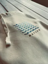 Hoodie de color crema con capucha y puños ajustados. Estampado trasero con un sol estilizado en color terracota y tres olas onduladas en color gris azulado.