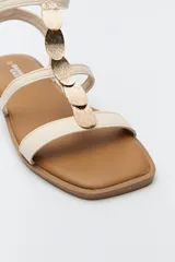 Sandalias planas color beige con tiras finas y aplique metálico central. Cierre con hebilla lateral y base marrón.