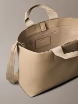 Cartera tote Calvin Klein color beige, de piel sintética granulada, con doble asa corta y correa larga ajustable con logo jacquard. Incluye cierre de correa magnético y bolsillos interiores.