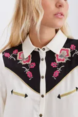 Camisa blanca de manga larga con canesú delantero y trasero en tela negra con bordados florales en tonos rosados y detalles en hilo dorado. Cierre frontal con botones metálicos tipo snap.