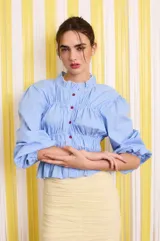 Camisa celeste a rayas verticales blancas, de corte entallado, con cuello clásico, cierre frontal con botones rojos, frunces en el delantero y mangas largas abullonadas con puños elásticos.