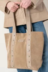 Bolso tipo tote de cuero vacuno gamuzado color verde oliva con apliques de tachas metálicas plateadas en los laterales. Tiene doble asa de cuero marrón y etiqueta de cuero con costuras en el frente.