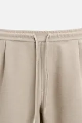 Bermuda color beige de corte recto, con cintura elástica ajustable con cordón, detalle de pliegues frontales, bolsillos laterales y bolsillos de vivo en la espalda.