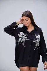 Camisa negra de manga larga con cuello clásico y botones frontales. Presenta dos grandes apliques bordados con lentejuelas y cuentas en forma de flores y hojas en la parte delantera.