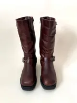 Bota de caña media de cuero vacuno color marrón oscuro, con punta cuadrada, cierre lateral y suela track negra. Presenta detalles de hebillas metálicas plateadas en la caña y en la parte inferior del empeine.