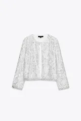 Chaqueta corta blanca con lentejuelas plateadas, cuello redondo, manga larga y forro interior.