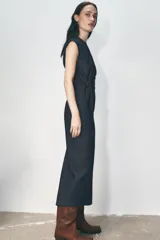 Vestido midi de denim azul oscuro, con cuello redondo y sisas. Presenta un aplique dorado en la cintura y abertura en la espalda. Cierre trasero con cremallera oculta.