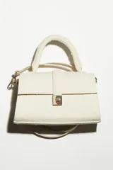 Cartera bandolera pequeña color crudo, de lona texturizada, con asa superior y correa ajustable extraíble. Tiene solapa con trabilla y cierre metálico dorado.