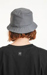 Gorro tipo bucket de color gris oscuro (carbón), con ala ancha y un pequeño parche negro con un logo bordado en blanco en el frente.