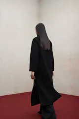 Abrigo largo de lana reciclada en color negro, con diseño minimalista, cuello redondo y cierre frontal con botones ocultos. Presenta aberturas laterales, bolsillos frontales tipo parche y mangas largas.