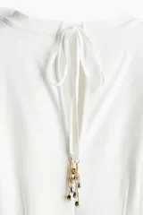 Remera larga color crudo, de punto pesado de algodón con cuello redondo y abertura en V en la espalda con tiras largas finas adornadas con dijes de cuentas brillantes. Modelo de hombros caídos con mangas amplias al codo y cintura entallada con pinzas adelante y atrás.