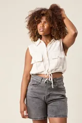 Chaleco cropped color crema, confeccionado en lino, con cuello camisero, cierre frontal con botones, dos bolsillos con solapa en el pecho y lazo ajustable en la cintura.