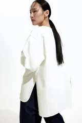 Blusa blanca de corte holgado en sarga de algodón con costuras visibles adelante y atrás. Modelo con cuello redondo, hombros caídos, tapeta con botones ocultos y mangas largas con puños abotonados. Bajo levemente redondeado con abertura pronunciada en los laterales. Espalda algo más larga.