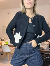Saco de lana negro, de corte clásico y abotonado, con cuello redondo. Presenta apliques de flores de crochet blancas en la parte delantera inferior. Los botones son redondos y plateados con un diseño grabado.