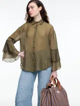 Camisa verde militar traslúcida con cuello clásico, cierre frontal con botones y mangas largas con volados. Presenta un broche dorado en el cuello.