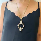 Collar con cadena de eslabones dorados y dije colgante de diseño geométrico en bronce. El dije presenta dos óvalos centrales de nácar y está rodeado por ocho cuentas pequeñas de perla o piedra blanca, engastadas en aros de bronce.