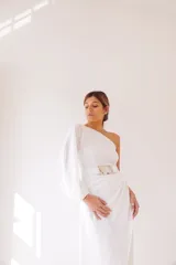 Vestido largo blanco de seda, con un hombro descubierto, manga larga abullonada y falda irregular con drapeado lateral. Incluye cinturón blanco con hebilla de alpaca y nácar.