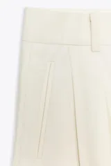 Pantalón fluido color crudo, de tiro medio con cintura con trabillas, bolsillos laterales, pinzas y raya planchada en delantero. Cierre frontal con cremallera, botón y ganchos metálicos.