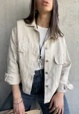 Sobrecamisa color beige con cuello camisero, cierre frontal con botones y bolsillos de parche en el pecho. Presenta detalles de broderie calado en mangas, hombros y bolsillos.