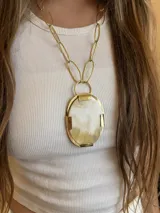 Collar con cadena de eslabones ovalados grandes en tono dorado, y un dije colgante grande de forma ovalada con borde dorado y centro de material claro con vetas marrones/beige.