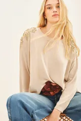 Sweater color beige con cuello redondo y mangas largas. Presenta apliques dorados en los hombros y un cinturón marrón con forma de flor.